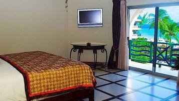 A hotel room with a bed, a bedside lamp, a television, a small table, and a view of a beach through a large window.