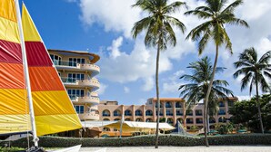 Un hotel frente a la playa con un velero, palmeras y un cielo despejado.