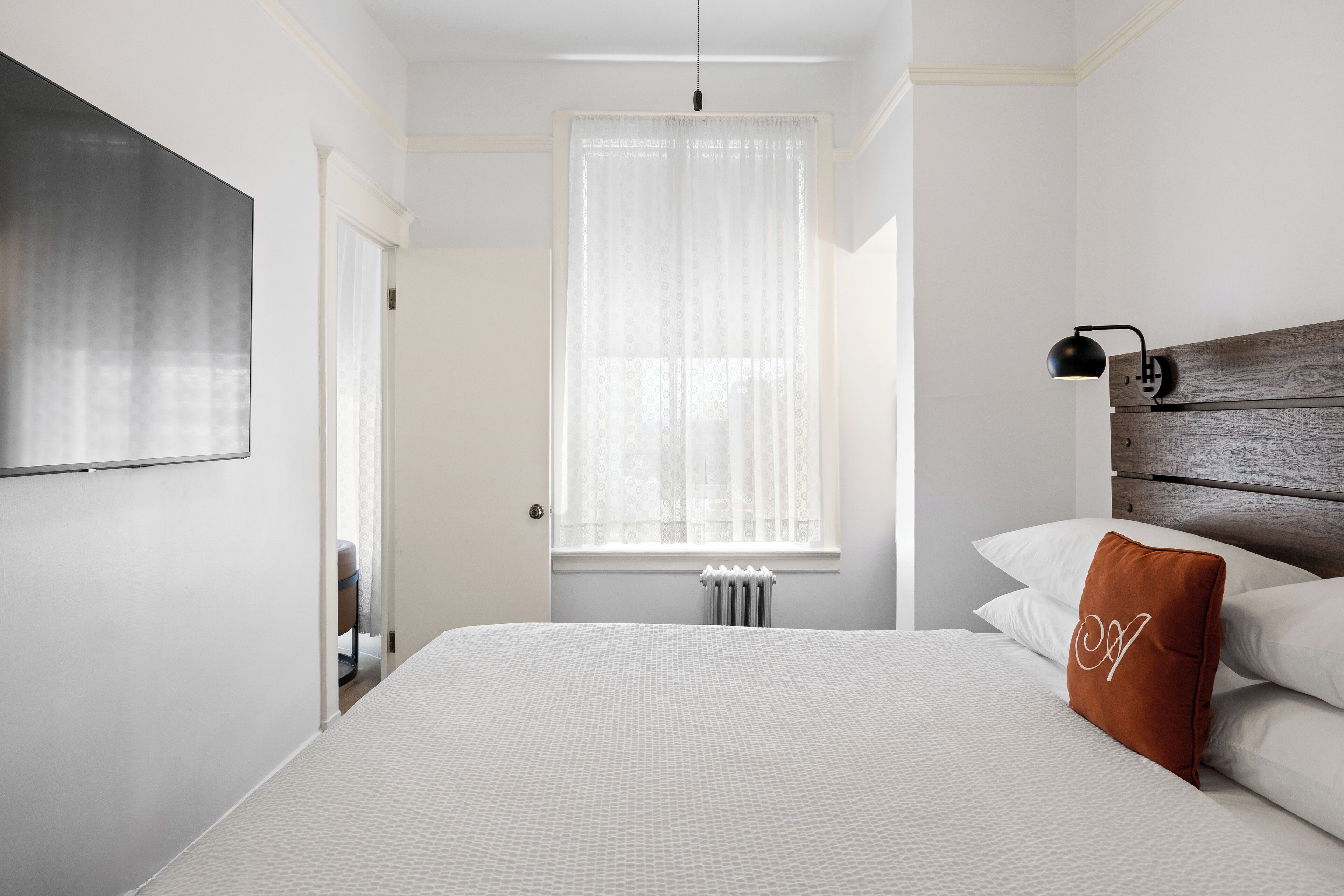 A bedroom with a large bed, a wooden headboard, a bedside lamp, and a window with sheer curtains.