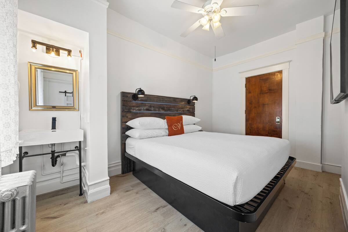 A bedroom with a bed, a wooden headboard, a white wall, a wooden door, and a ceiling fan.