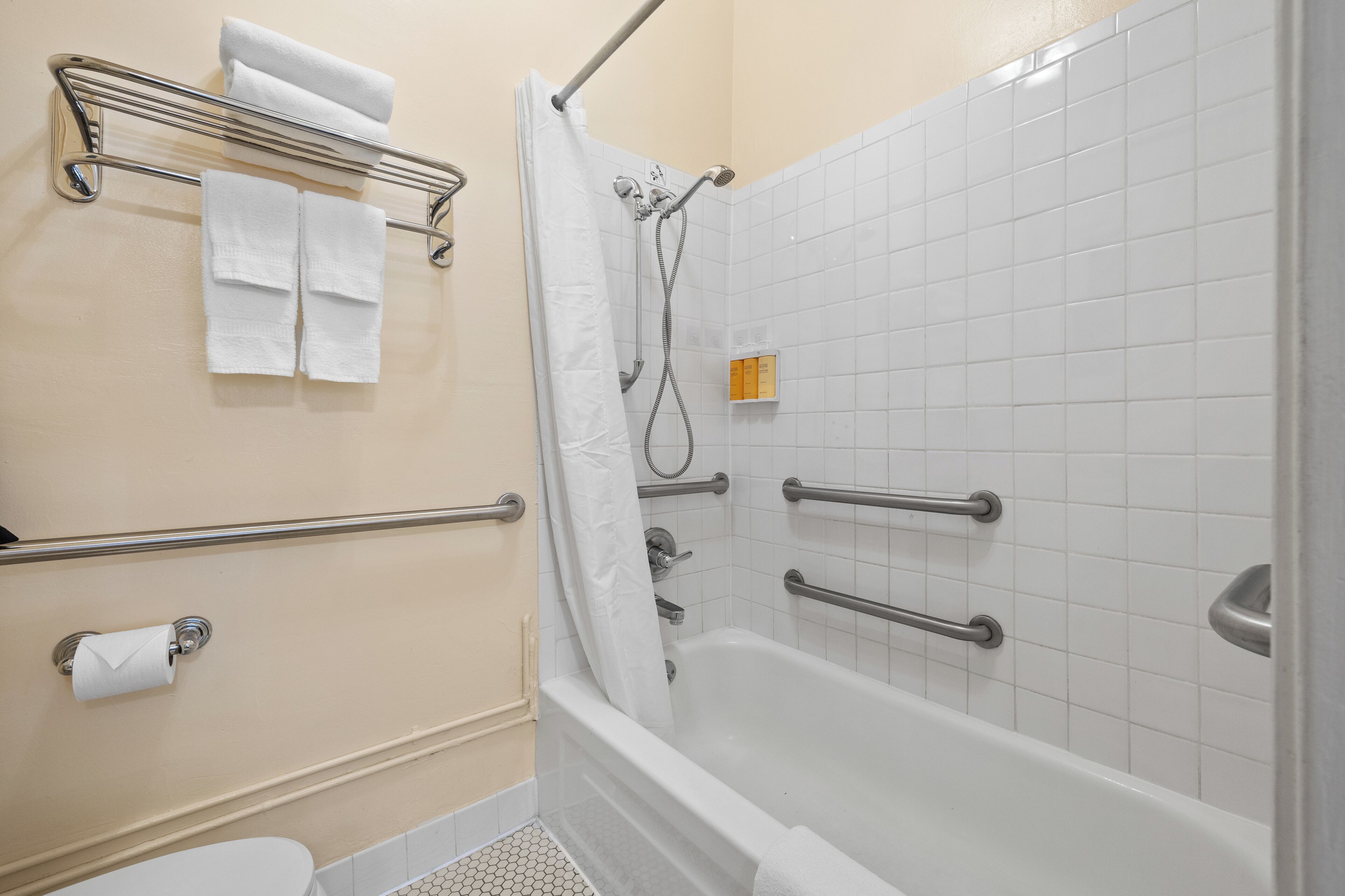 Un baño con bañera, ducha e inodoro. Hay barras de apoyo y un perchero para toallas.