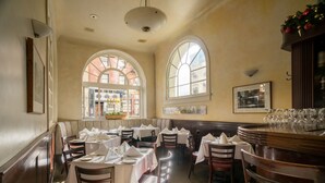 Ein Restaurant mit runden Tischen, weißen Tischdecken und Holzsesseln. Große Fenster mit Rundbögen und ein Selbstbedienungsbereich mit einer Theke und Hockern.