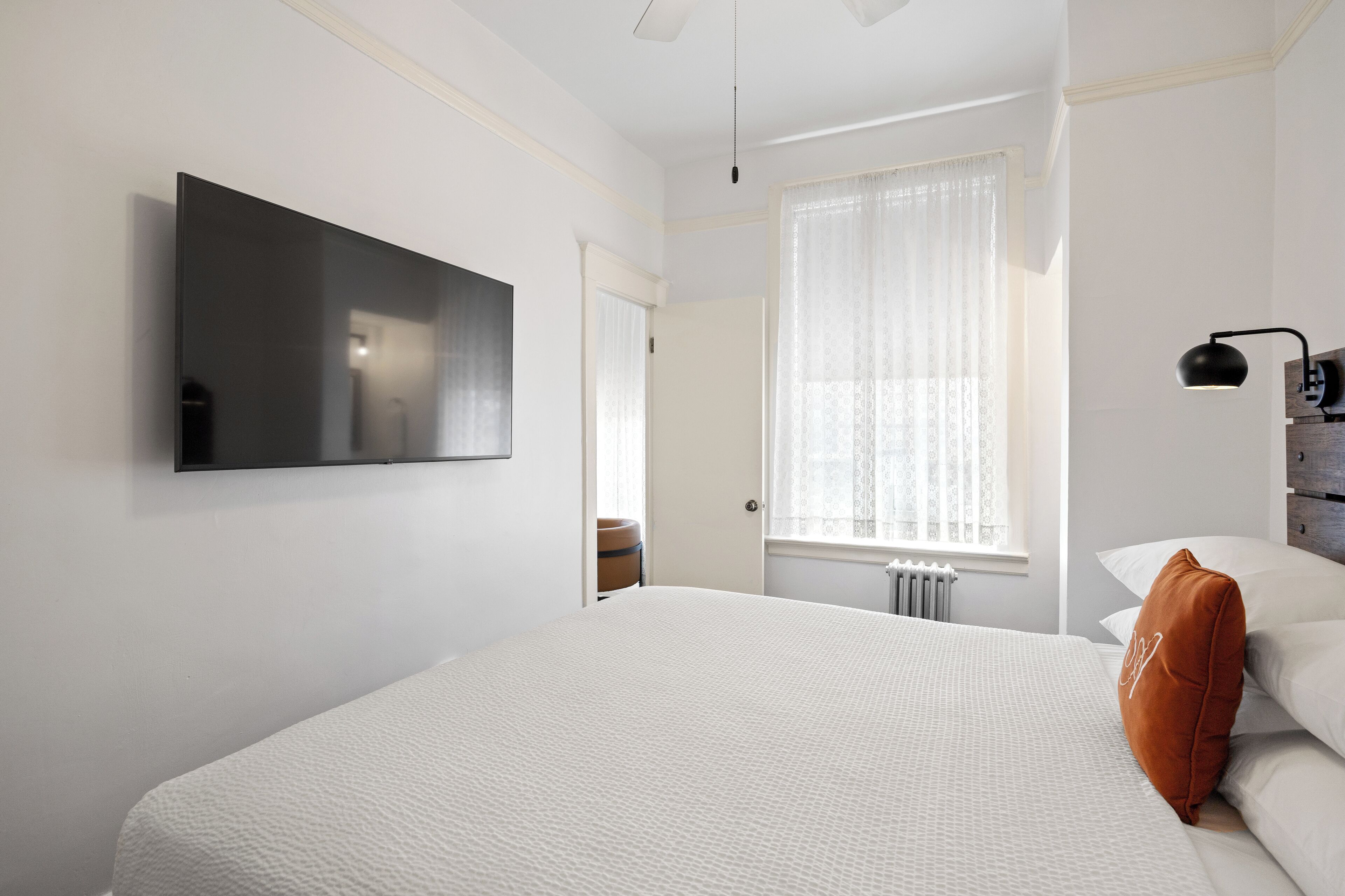 A bedroom with a large bed, a flat-screen TV mounted on the wall, a window with sheer curtains, and a bedside lamp.