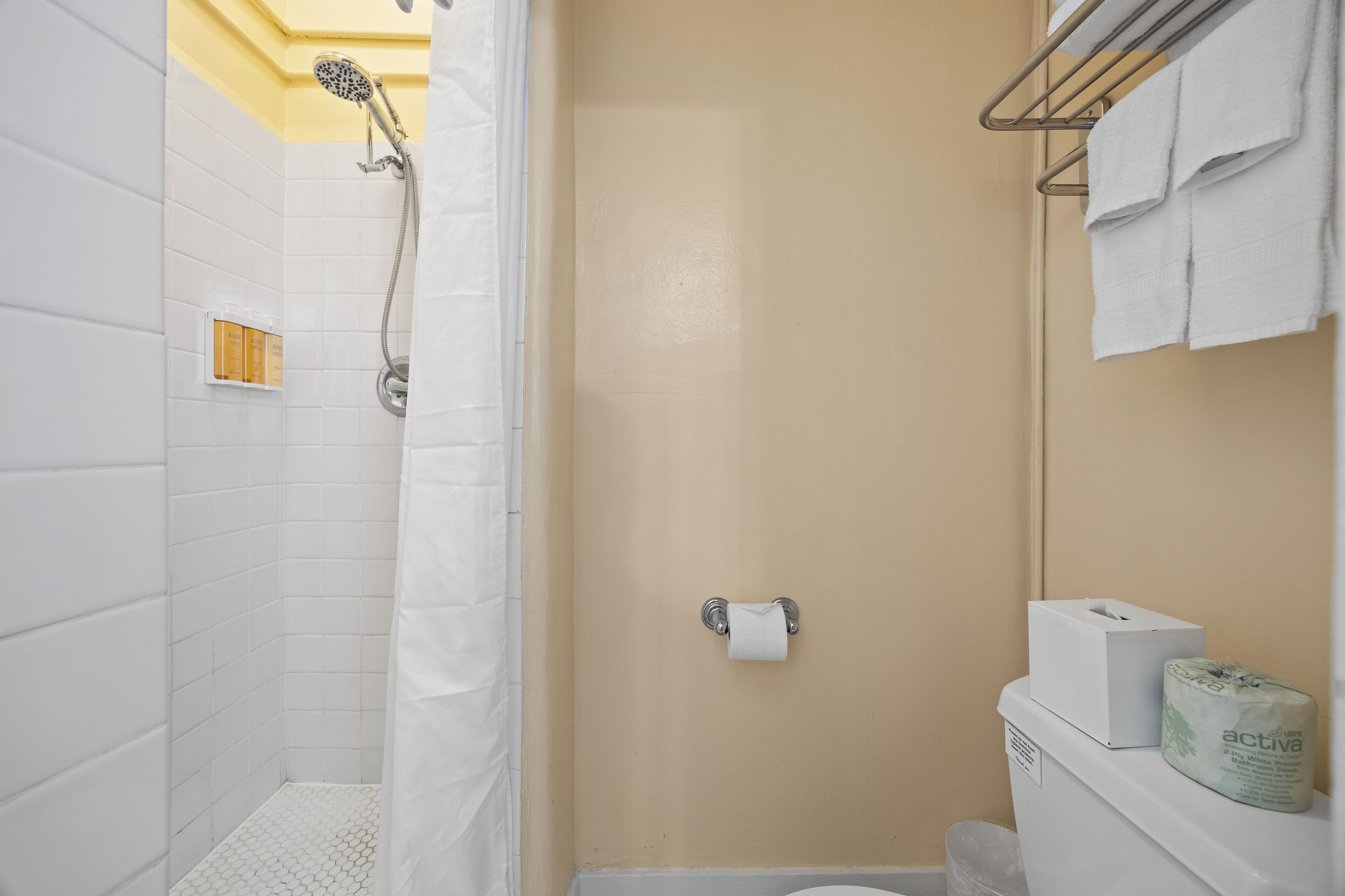 Un baño con paredes de azulejos blancos, una ducha con cabezal de ducha manual, un inodoro con dispensador de toallas de papel y papel higiénico, y un perchero con toallas.