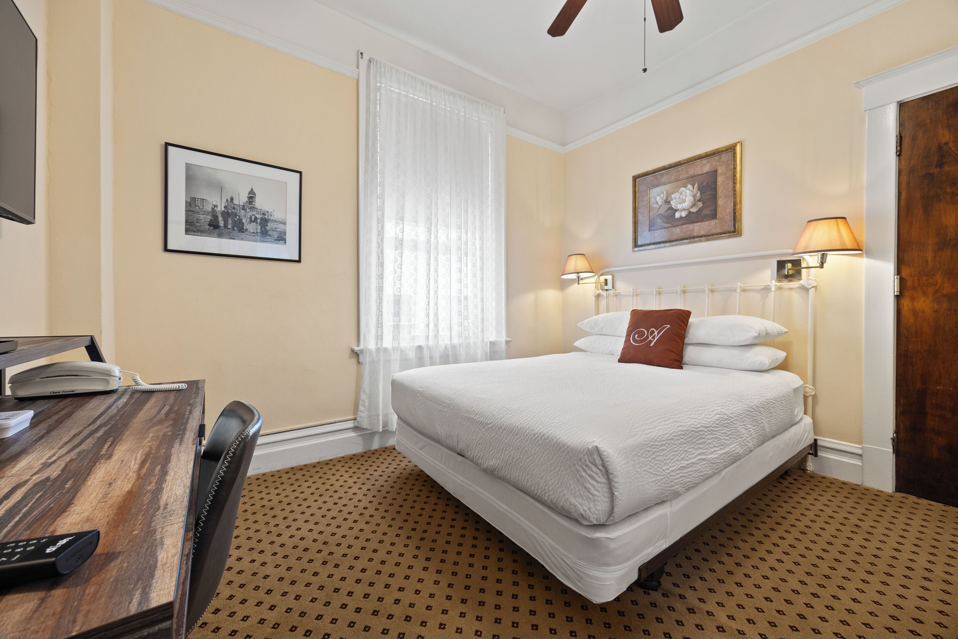 Une chambre d’hôtel comprenant un lit, un bureau avec un téléphone, une chaise, une fenêtre avec des rideaux et des lampes fixées au mur.