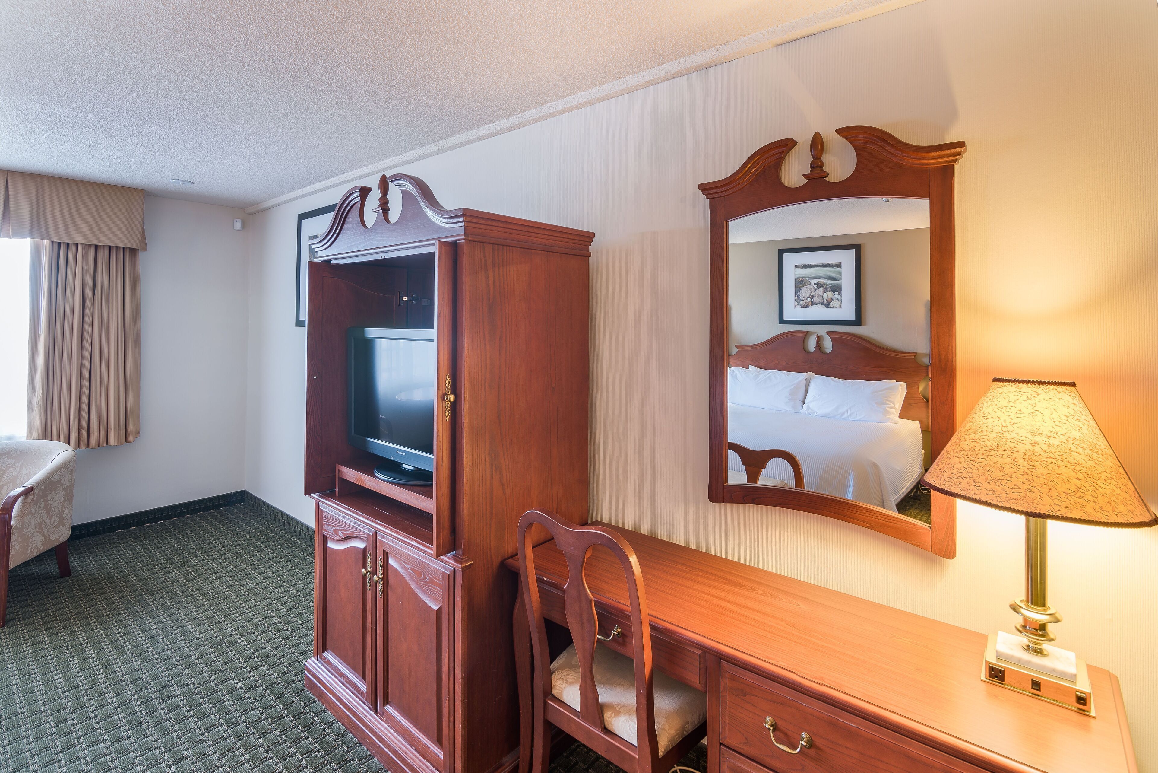 Een hotelkamer met een houten bureau, een televisie, een spiegel, een lamp en een bed.