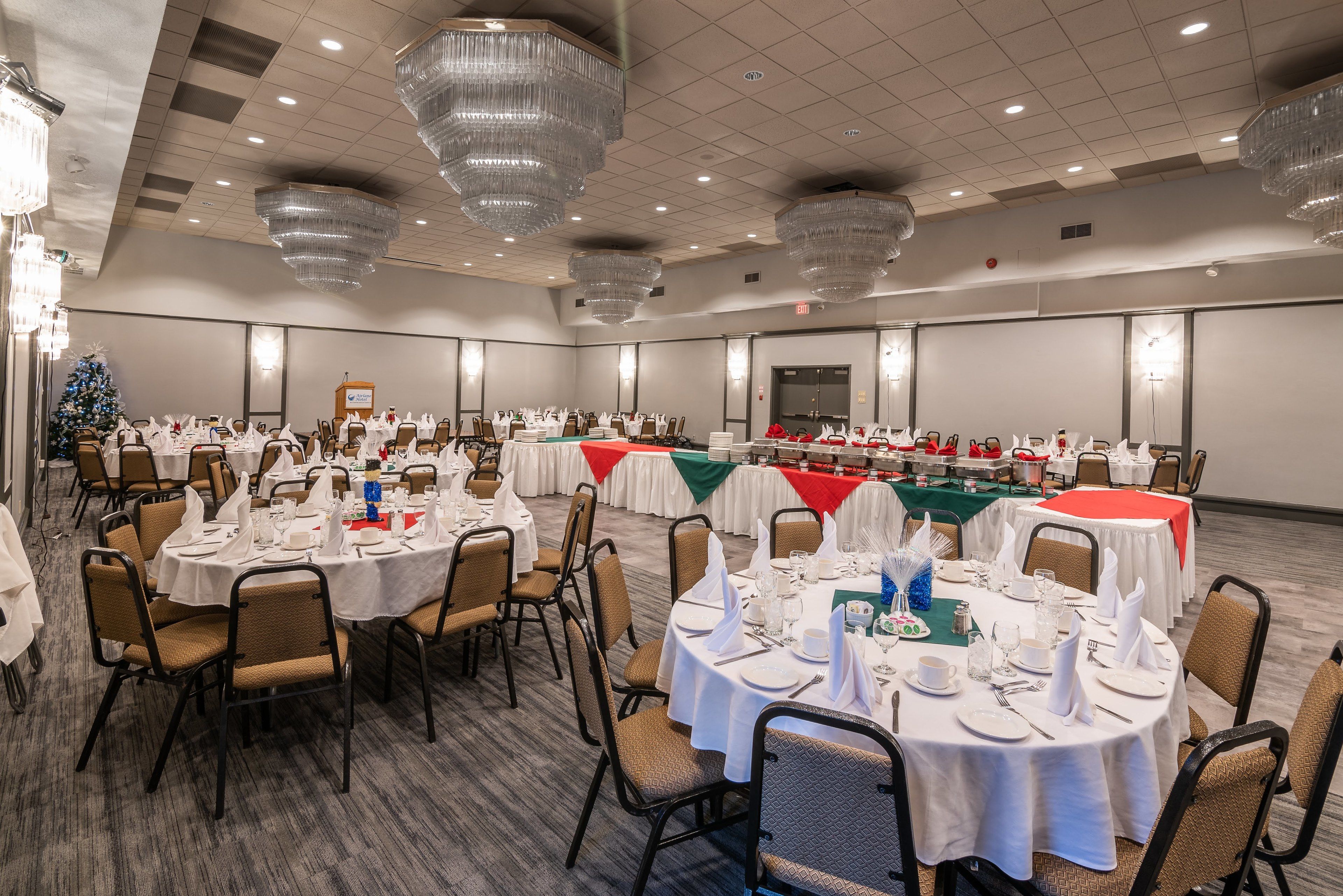 Un salón de banquetes con mesas redondas dispuestas para comer, con manteles blancos y sillas, y un árbol de Navidad al fondo.