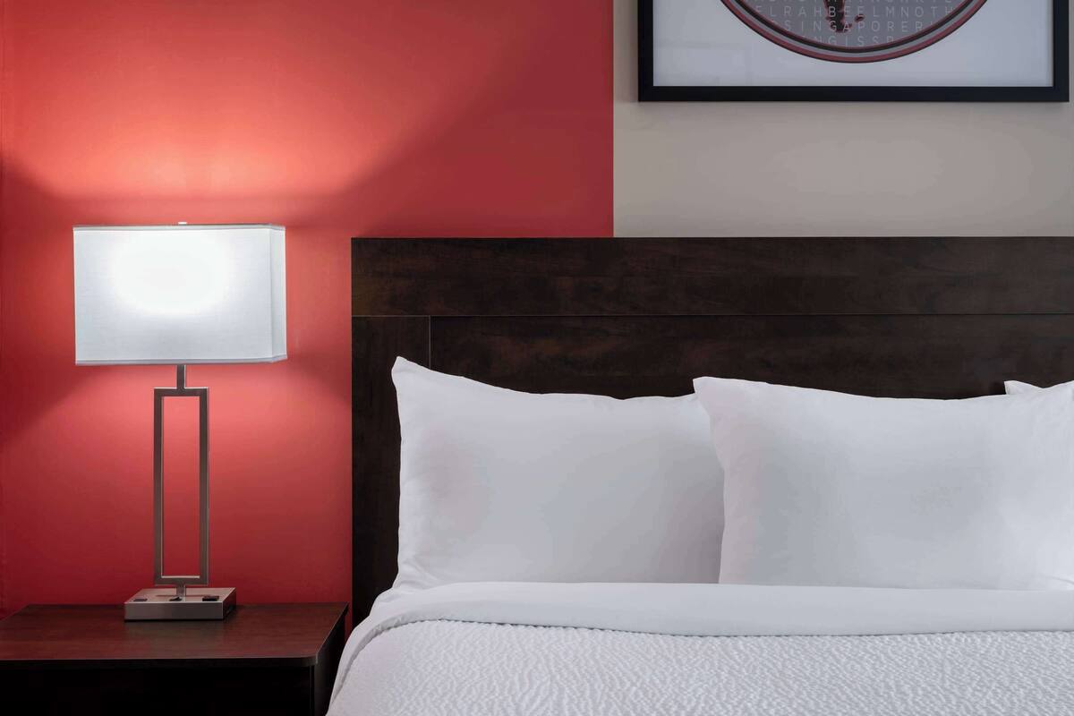 A neatly made bed with white bedding and pillows, a modern bedside table with a lamp, and a framed artwork on the wall.