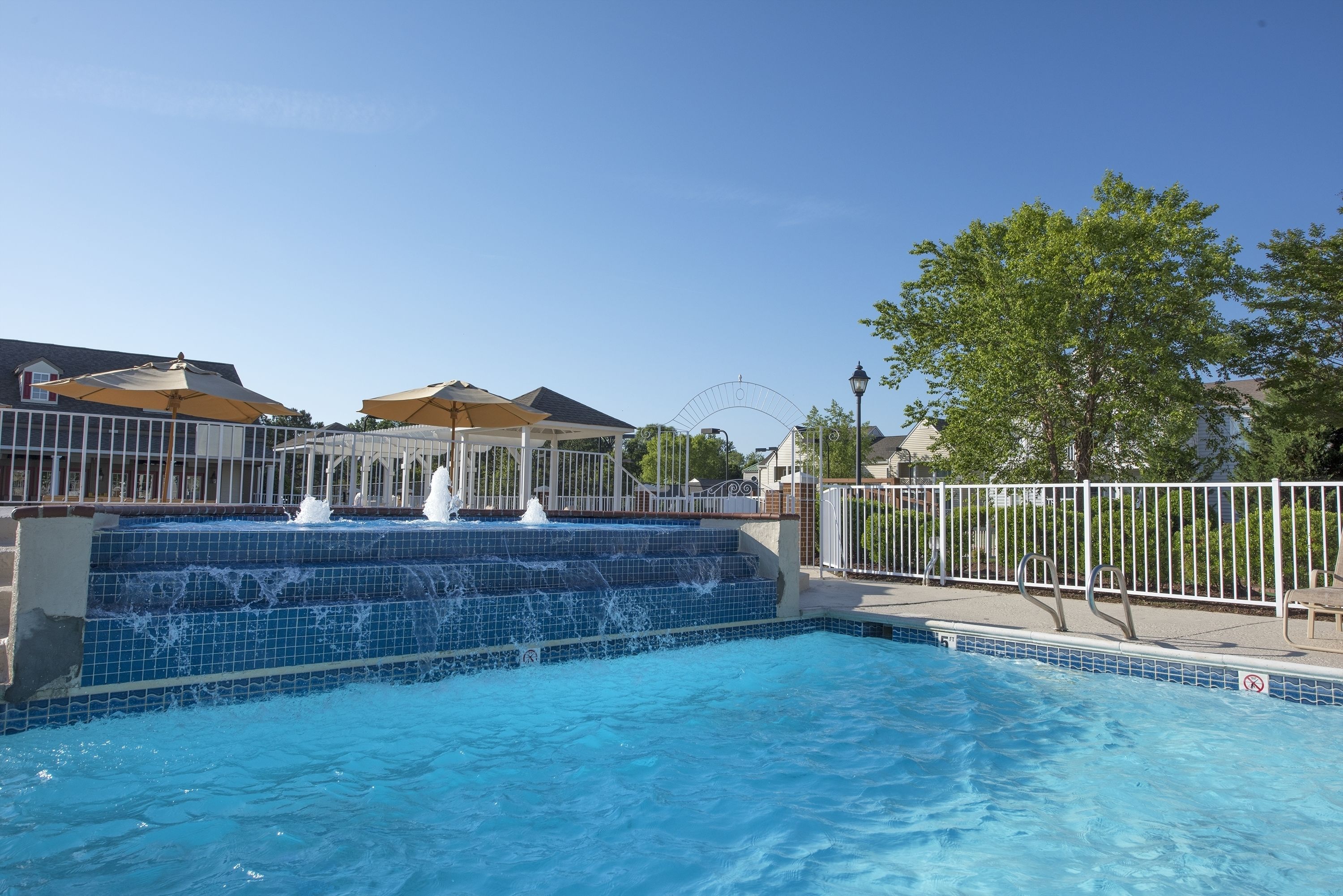 A swimming pool with a water feature, surrounded by a fence and umbrellas.