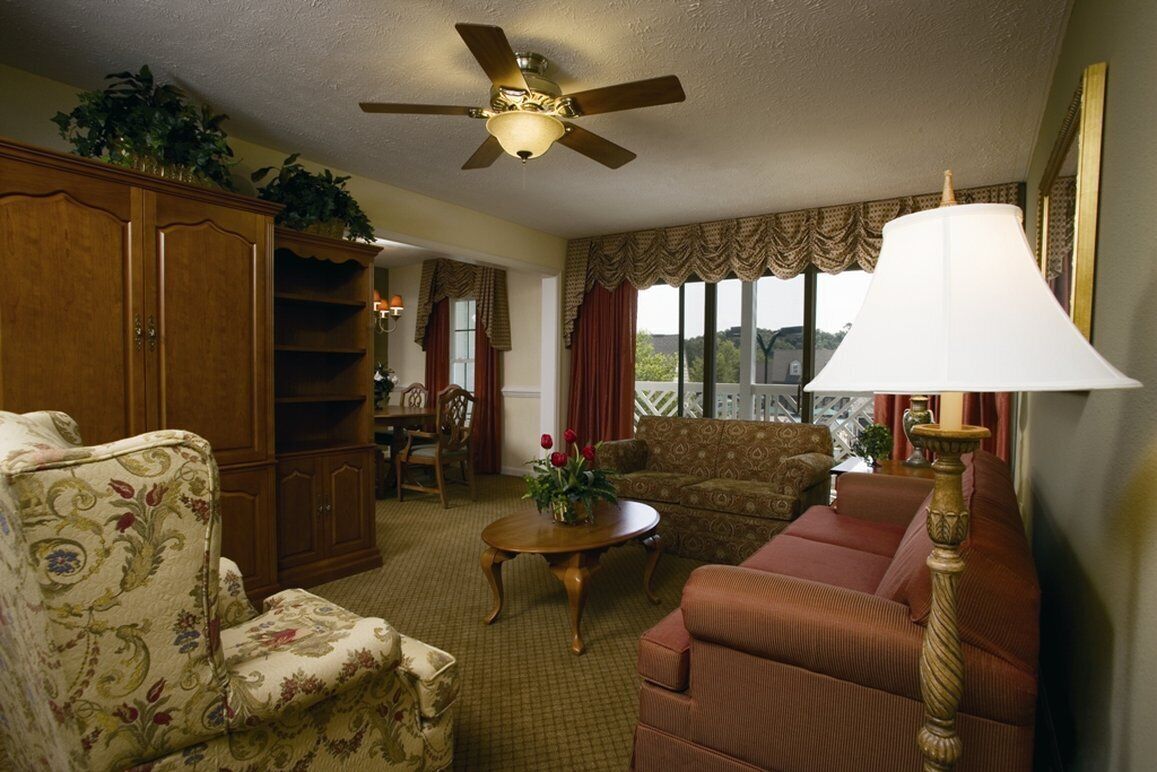 Un salon avec un ventilateur de plafond, un canapé, un fauteuil, une table ornée de fleurs et une lampe.