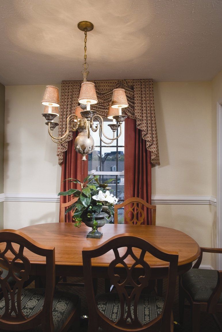 A dining area with a wooden table, six chairs, a chandelier, and a window with curtains.