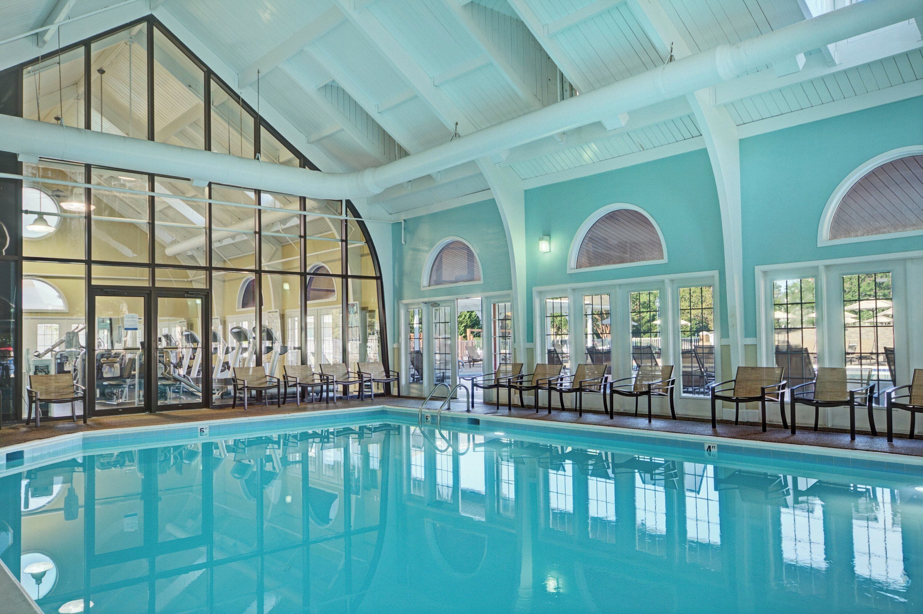 Una amplia piscina interior con paredes de vidrio transparente, múltiples sillones de descanso y un techo alto con vigas a la vista.