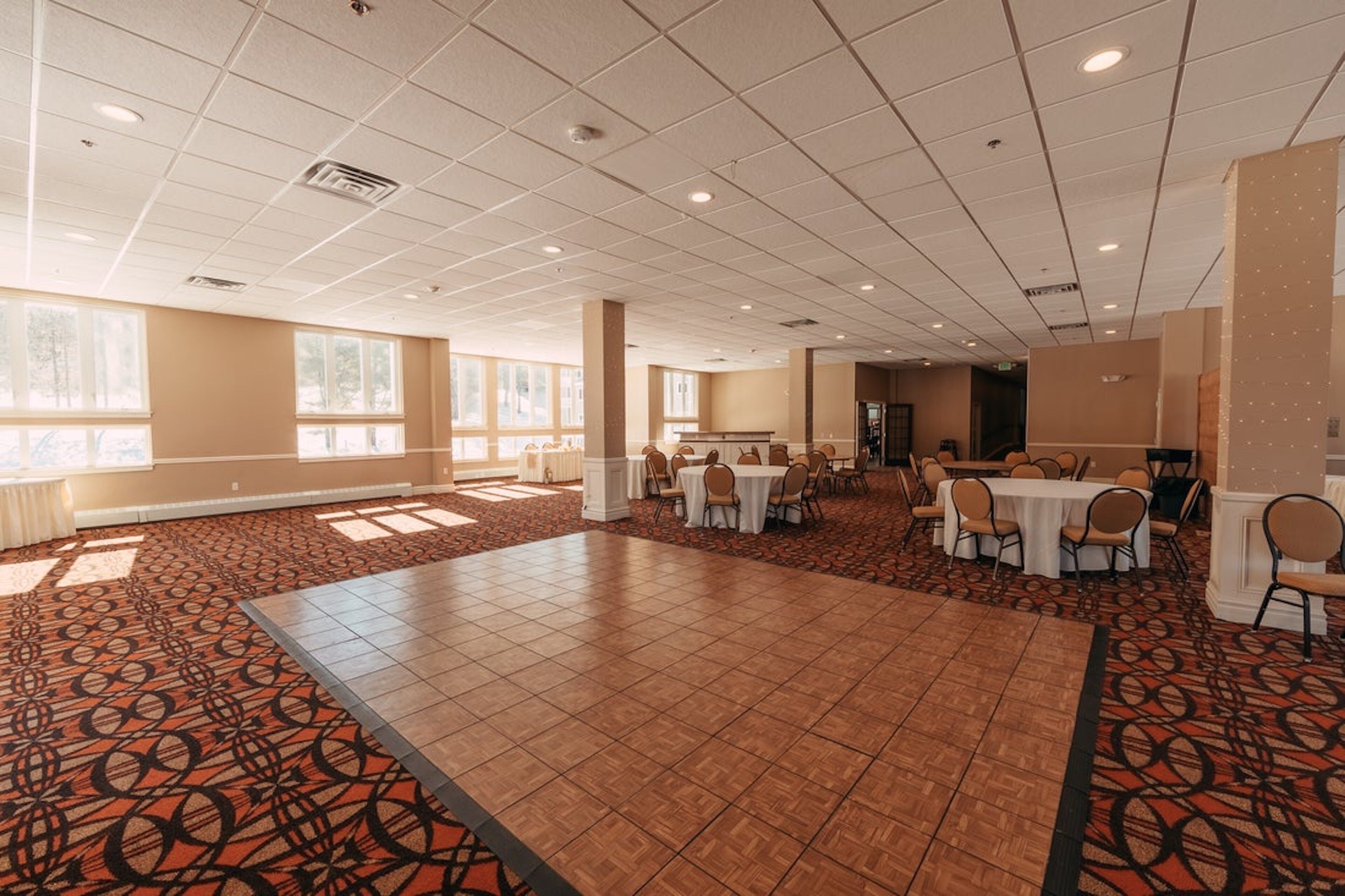 A spacious hall with a patterned carpet, multiple tables with chairs, and large windows allowing natural light.