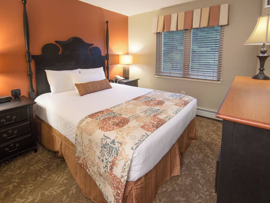 A hotel room with a four-poster bed, bedside tables with lamps, a dresser, and a window with blinds.