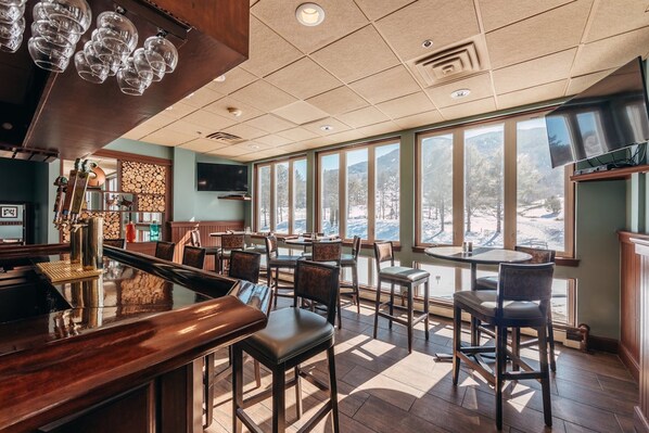 A bar area with a wooden counter and stools, large windows with a view of a snowy landscape, and a ceiling with recessed lighting.