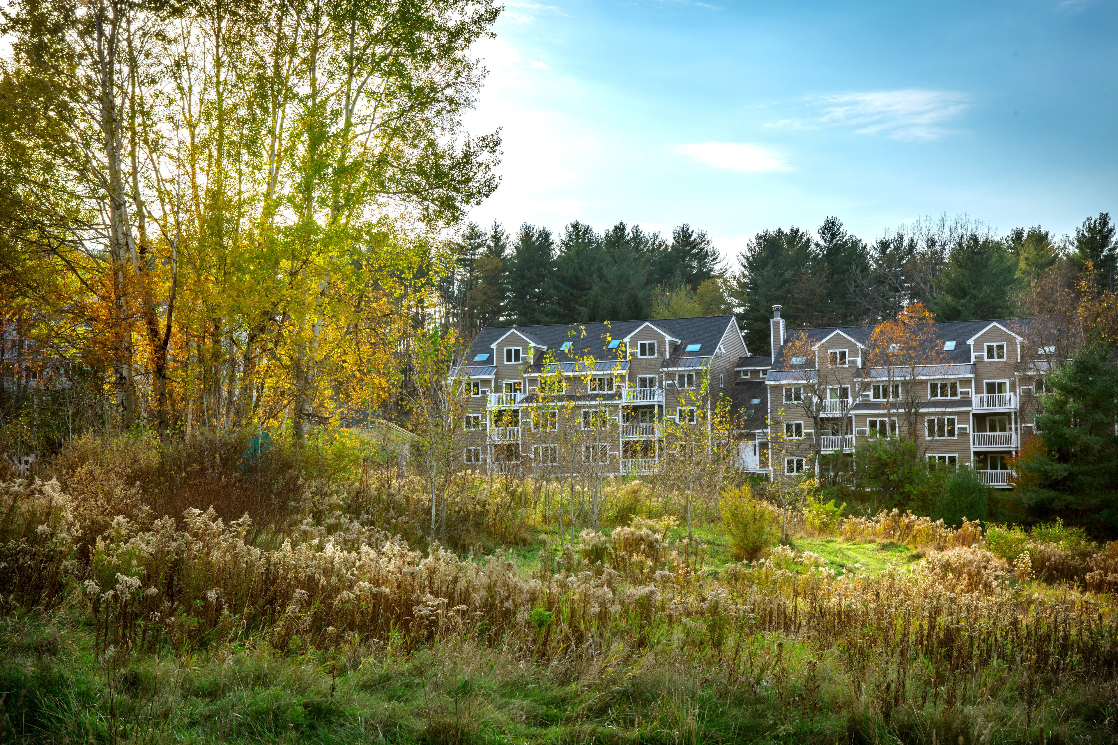 A residential building with multiple balconies, surrounded by autumn foliage and grassy areas.