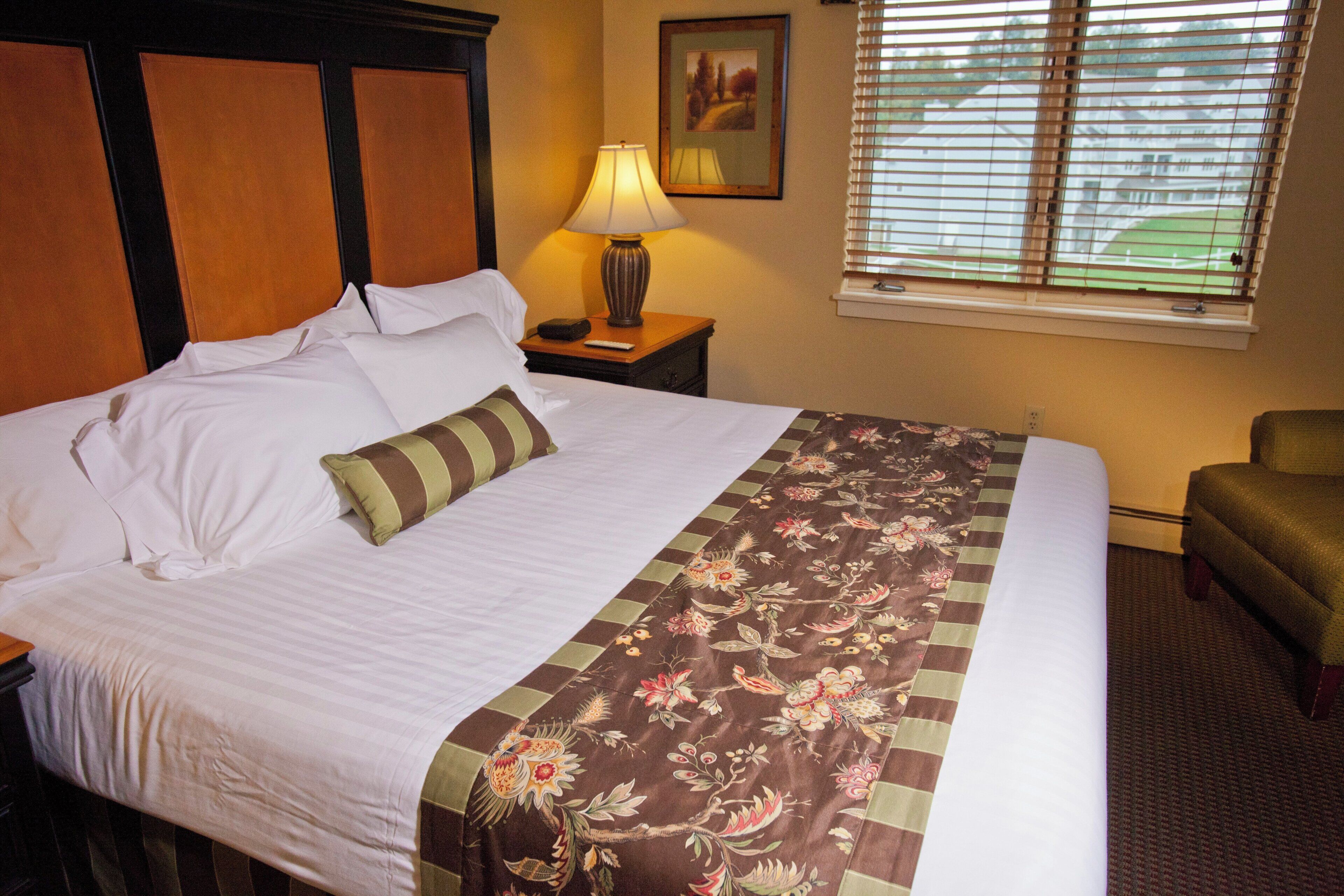 A hotel room with a bed, a nightstand, a lamp, a chair, and a window with blinds.
