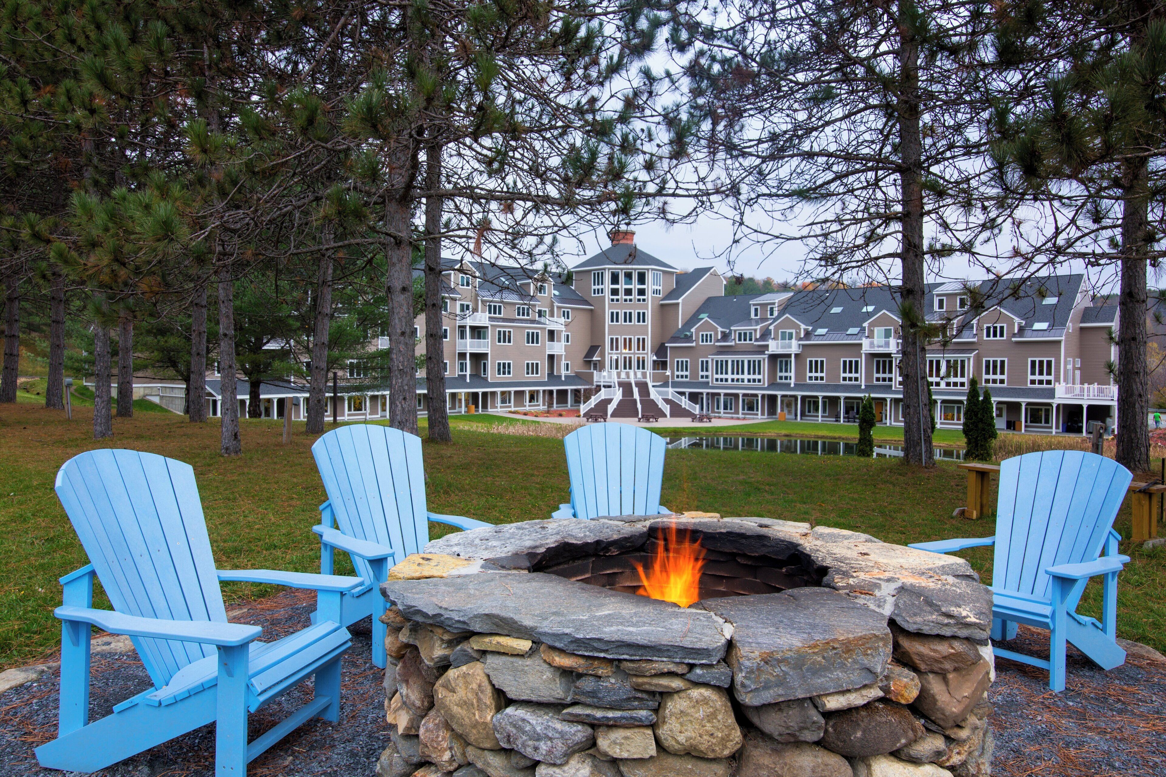 A stone fire pit with chairs around it, set in a grassy area with trees and a large building in the background.