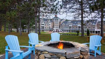 A stone fire pit with chairs around it, set in a grassy area with trees and a large building in the background.