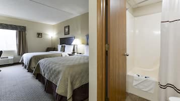 Habitación de hotel con dos camas, un escritorio, una silla, una lámpara y baño con bañera y cortina de ducha.