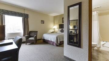 A hotel room with a bed, a chair, a desk, a lamp, a mirror, and a bathroom with a shower curtain.