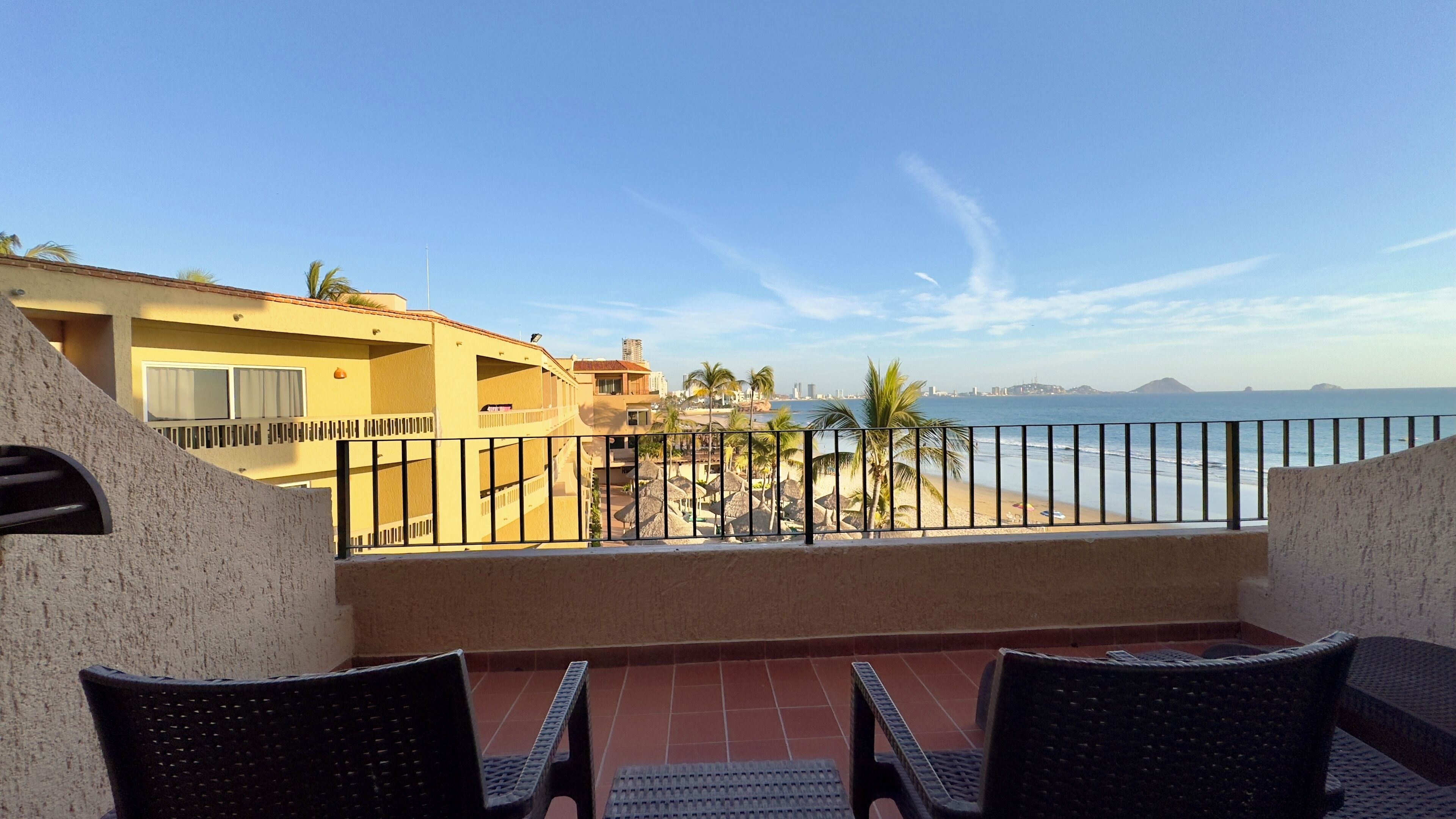 A balcony with two wicker chairs, a view of a beach, palm trees, and a cityscape in the distance.