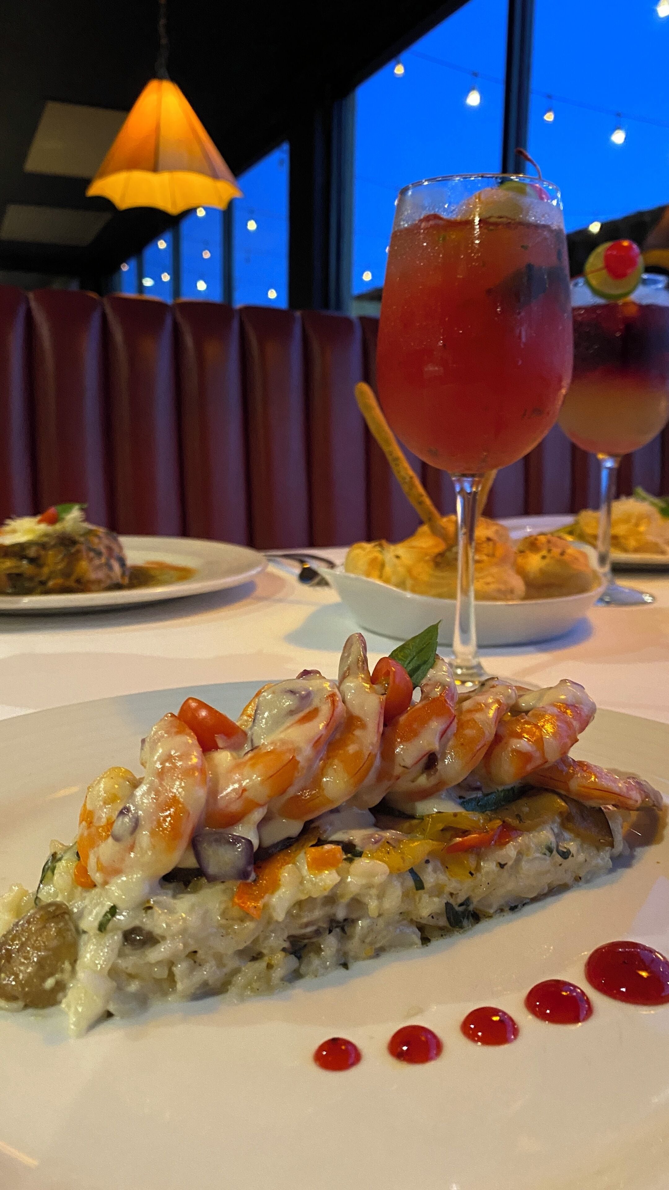 Una mesa de comedor con un plato de pasta de mariscos, un vaso de bebida roja y un postre con una cereza en un palito.