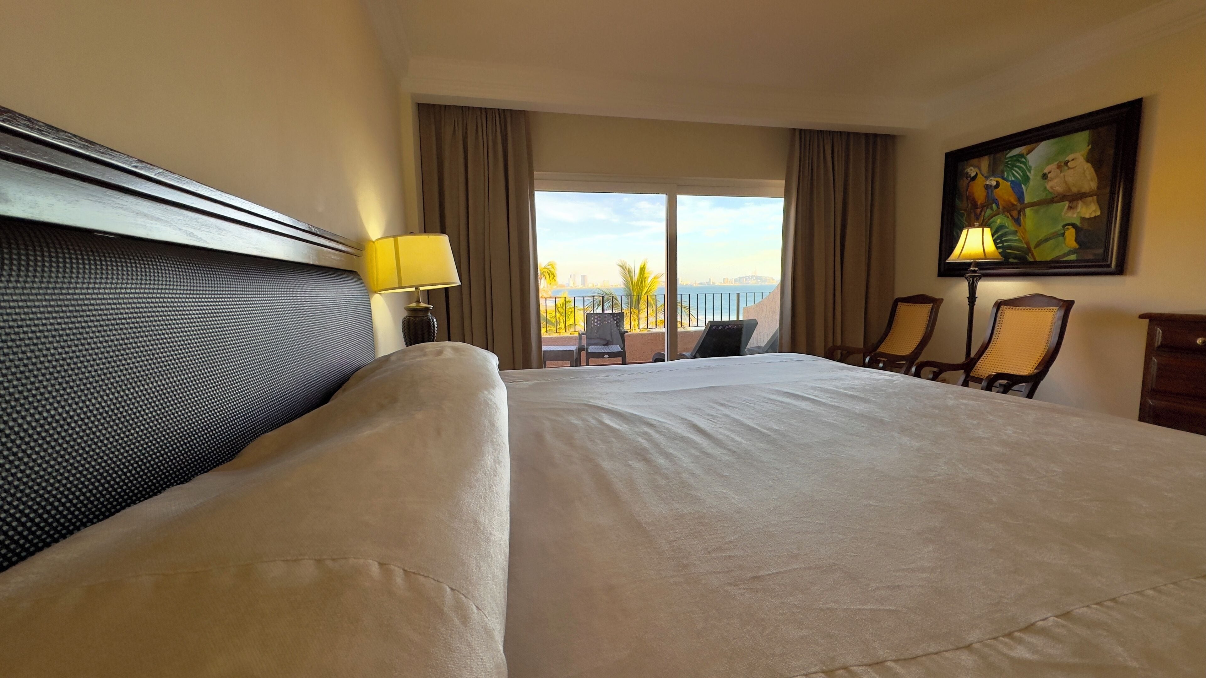 A hotel room with a large bed, a desk, two chairs, a painting, and a view of the city and palm trees.