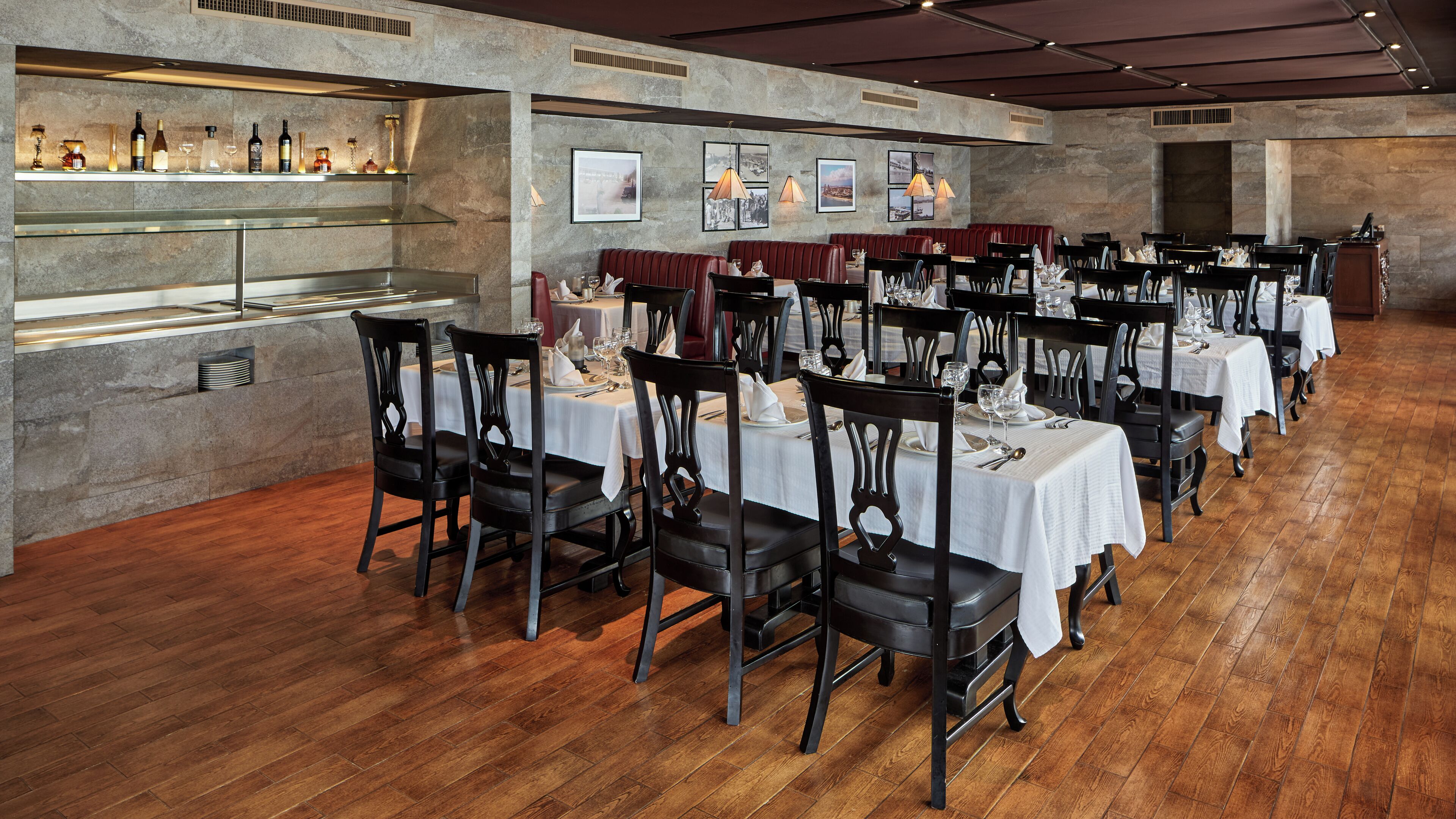 A restaurant with long wooden tables, black chairs, and a bar area with bottles and glasses.