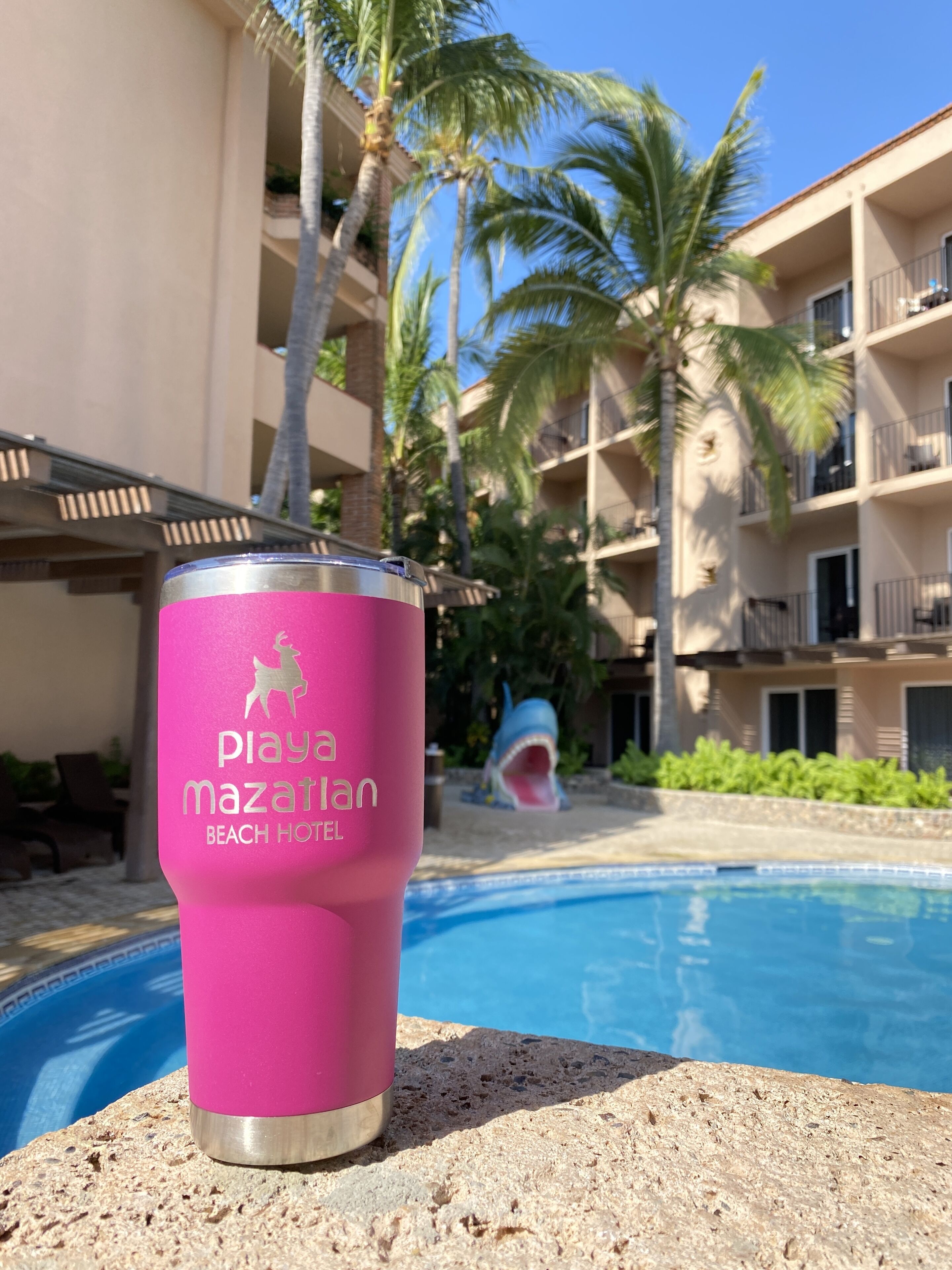 Un gobelet rose orné d’un logo, posé sur une surface en pierre, près d’une piscine, avec un bâtiment en arrière-plan.
