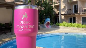 A pink tumbler with a logo, placed on a stone surface near a pool with a building in the background.