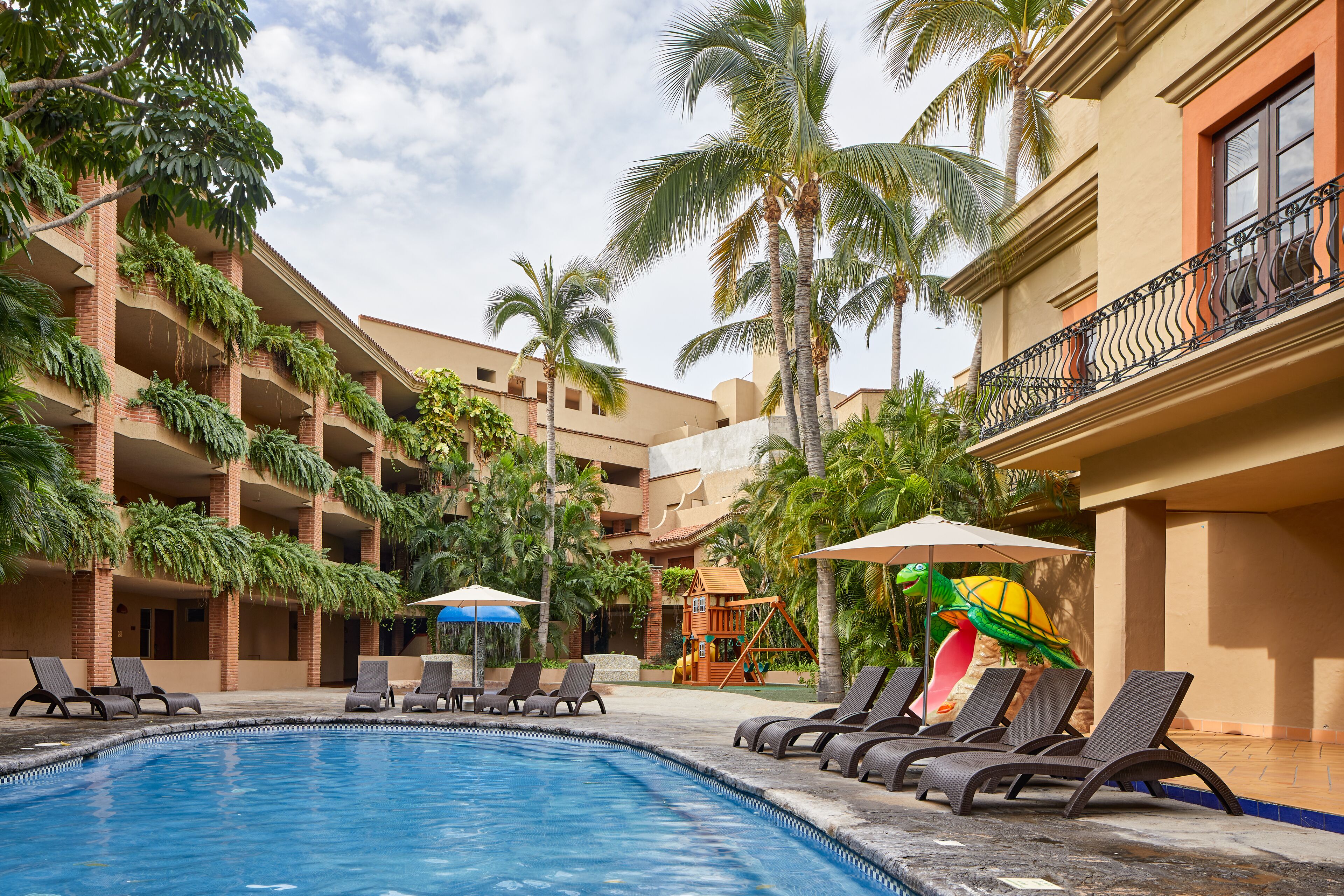A pool area with lounge chairs, palm trees, and a children's play structure.