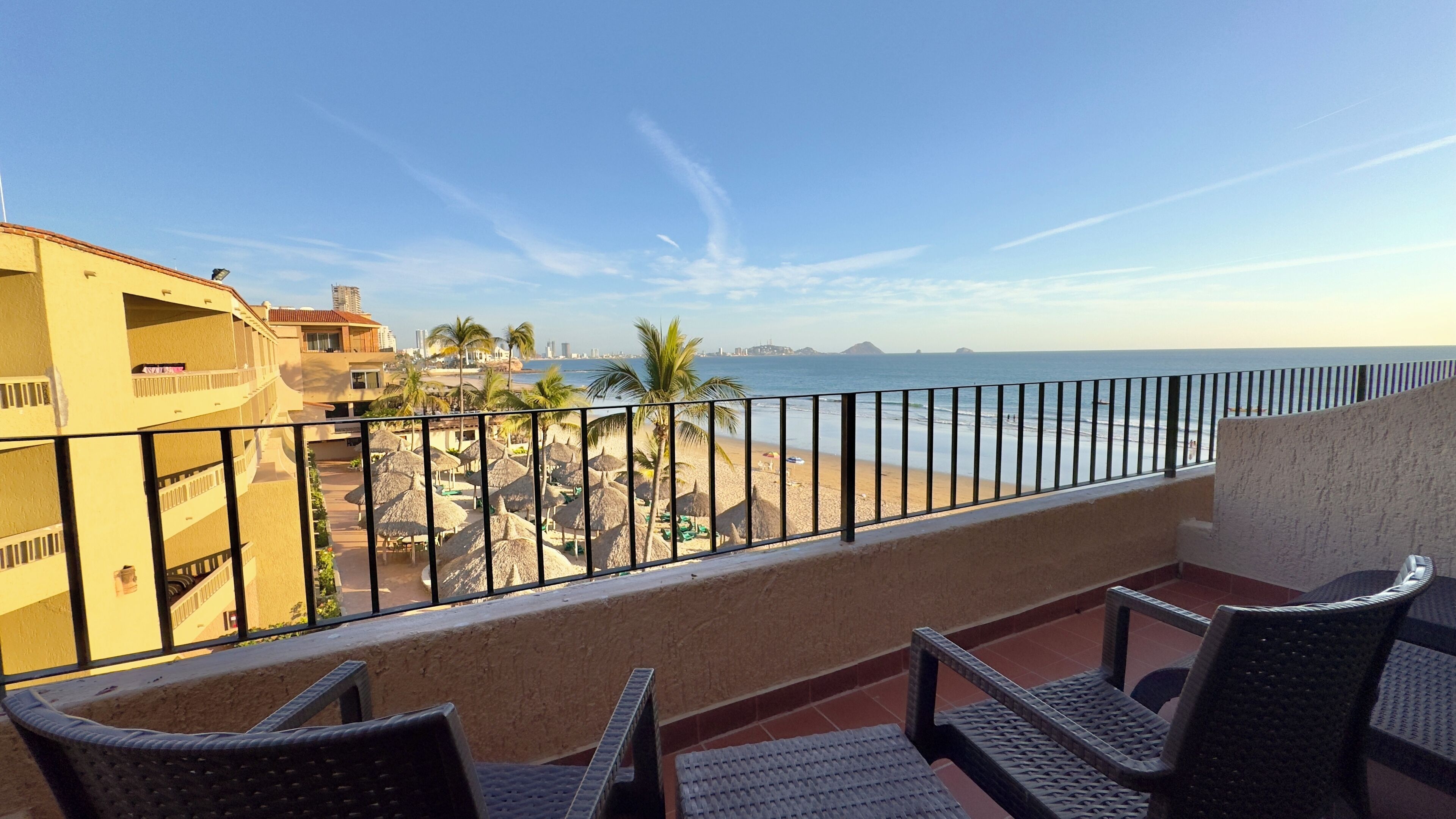 A balcony with wicker chairs, a view of a beach with palm trees, and a cityscape in the background.