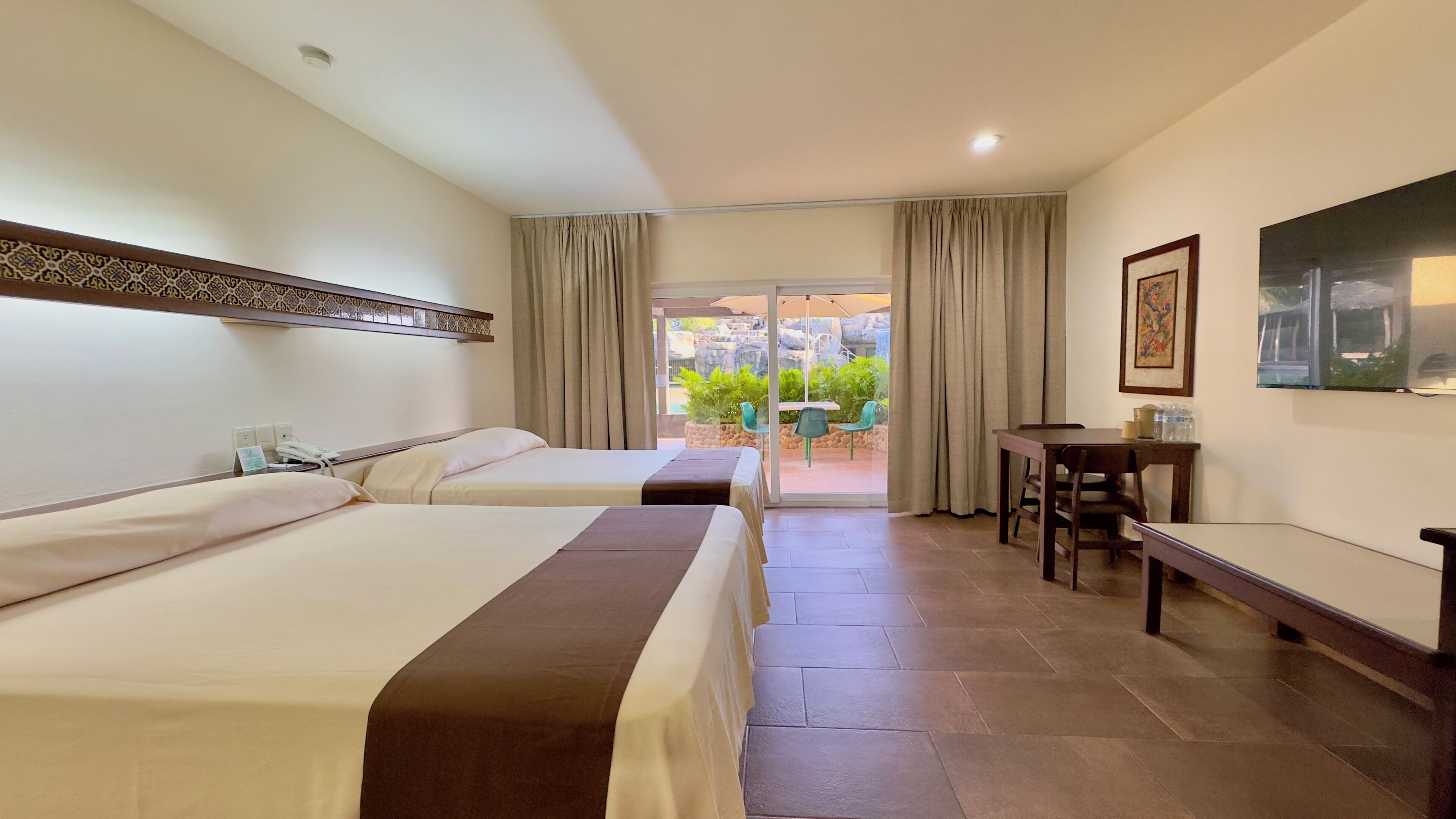 Quarto de hotel com duas camas, uma escrivaninha, uma cadeira e vista para área externa com vegetação e piscina.