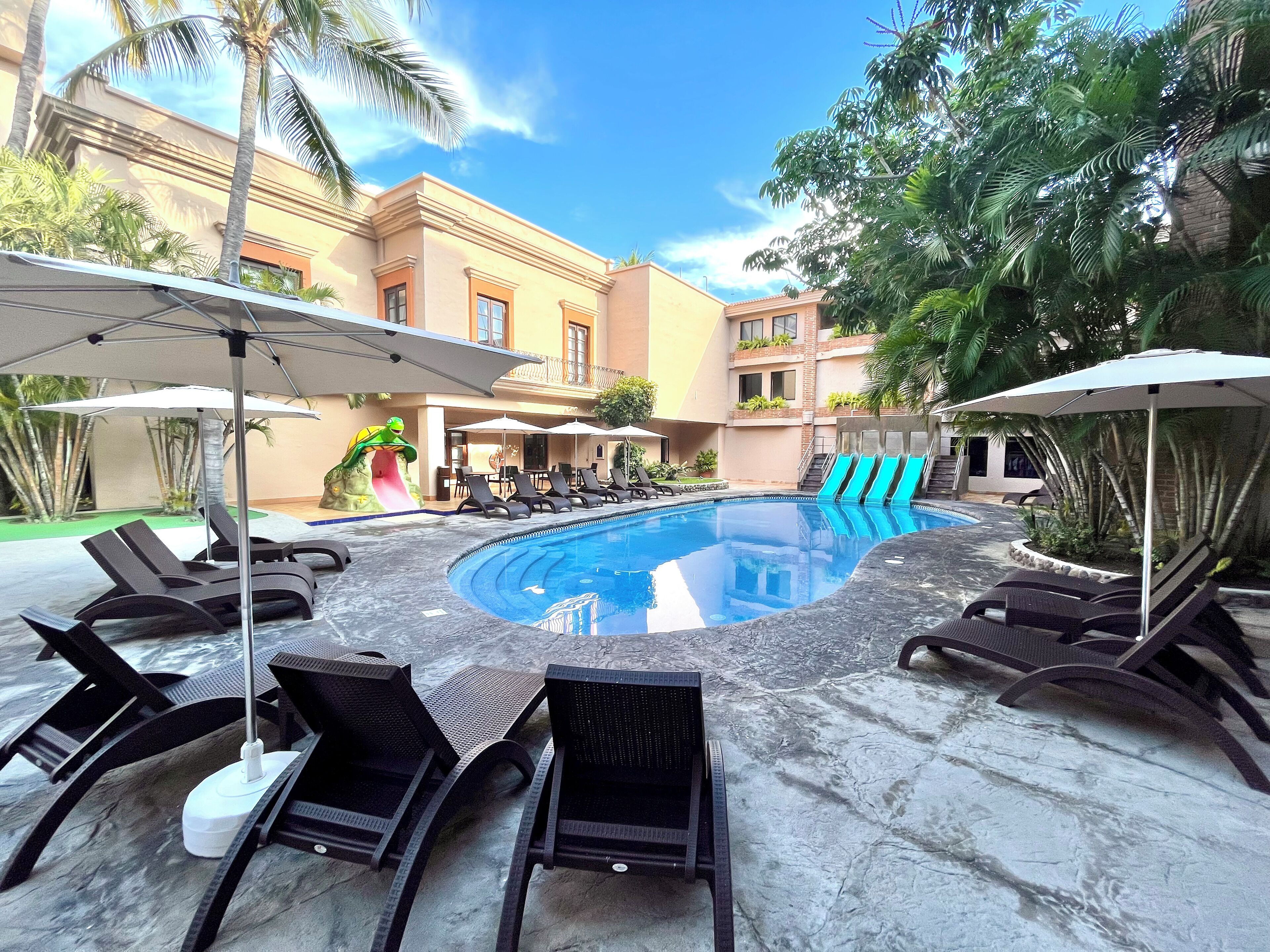 A pool area with lounge chairs and umbrellas, surrounded by buildings and palm trees.