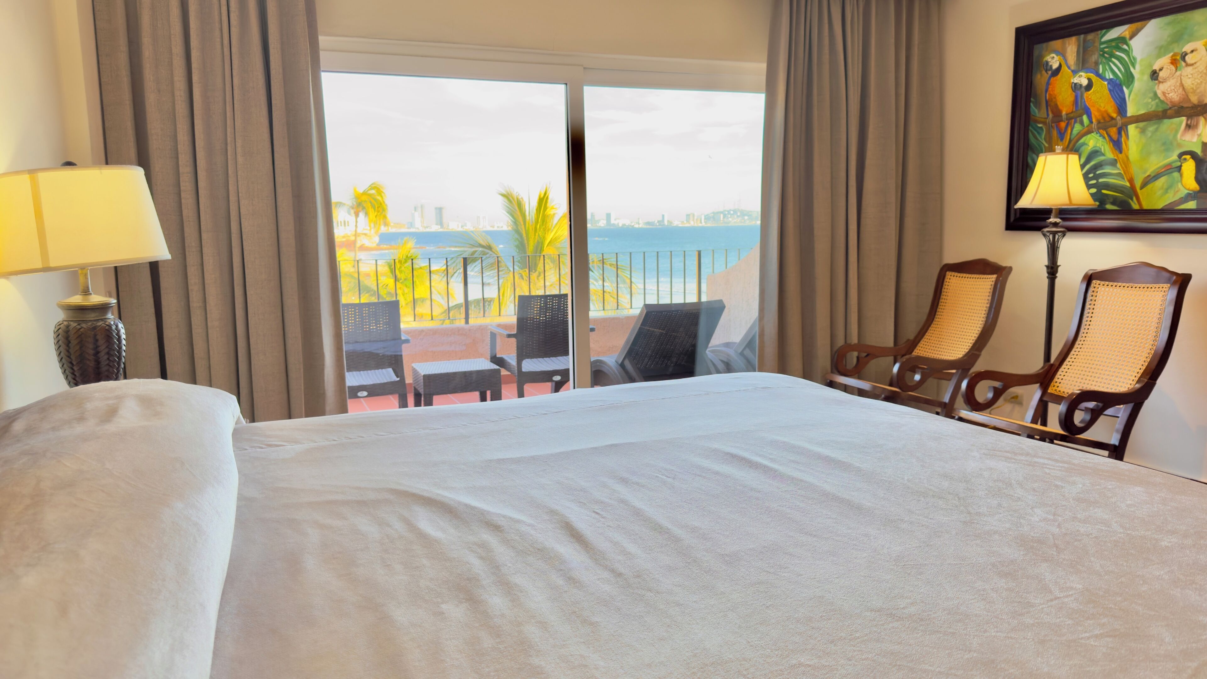 A hotel room with a large bed, a balcony with chairs, a painting, and a lamp.