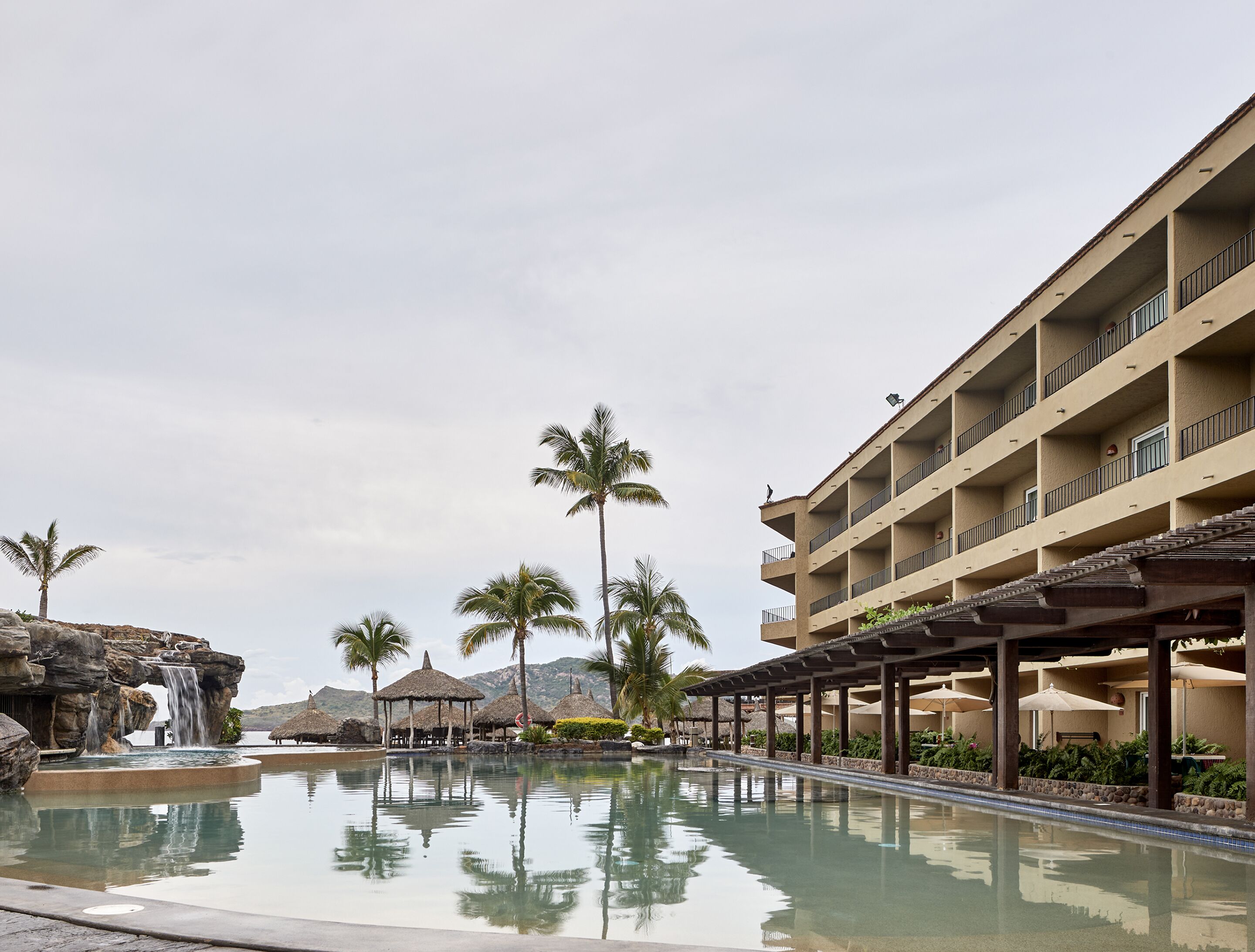 A resort pool with a waterfall, palm trees, and a multi-story building with balconies.