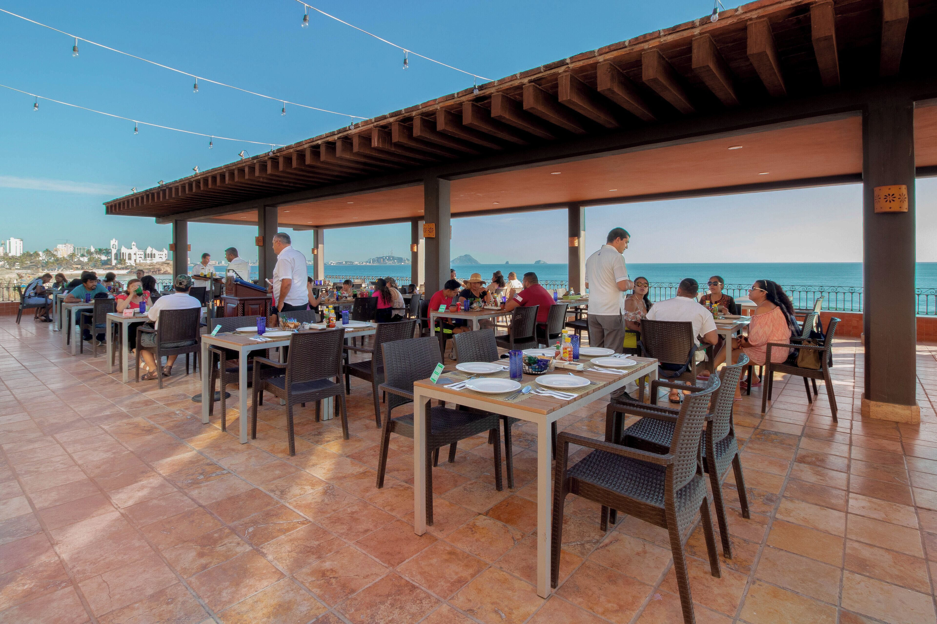 An outdoor dining area with tables and chairs, overlooking a body of water and a distant cityscape.