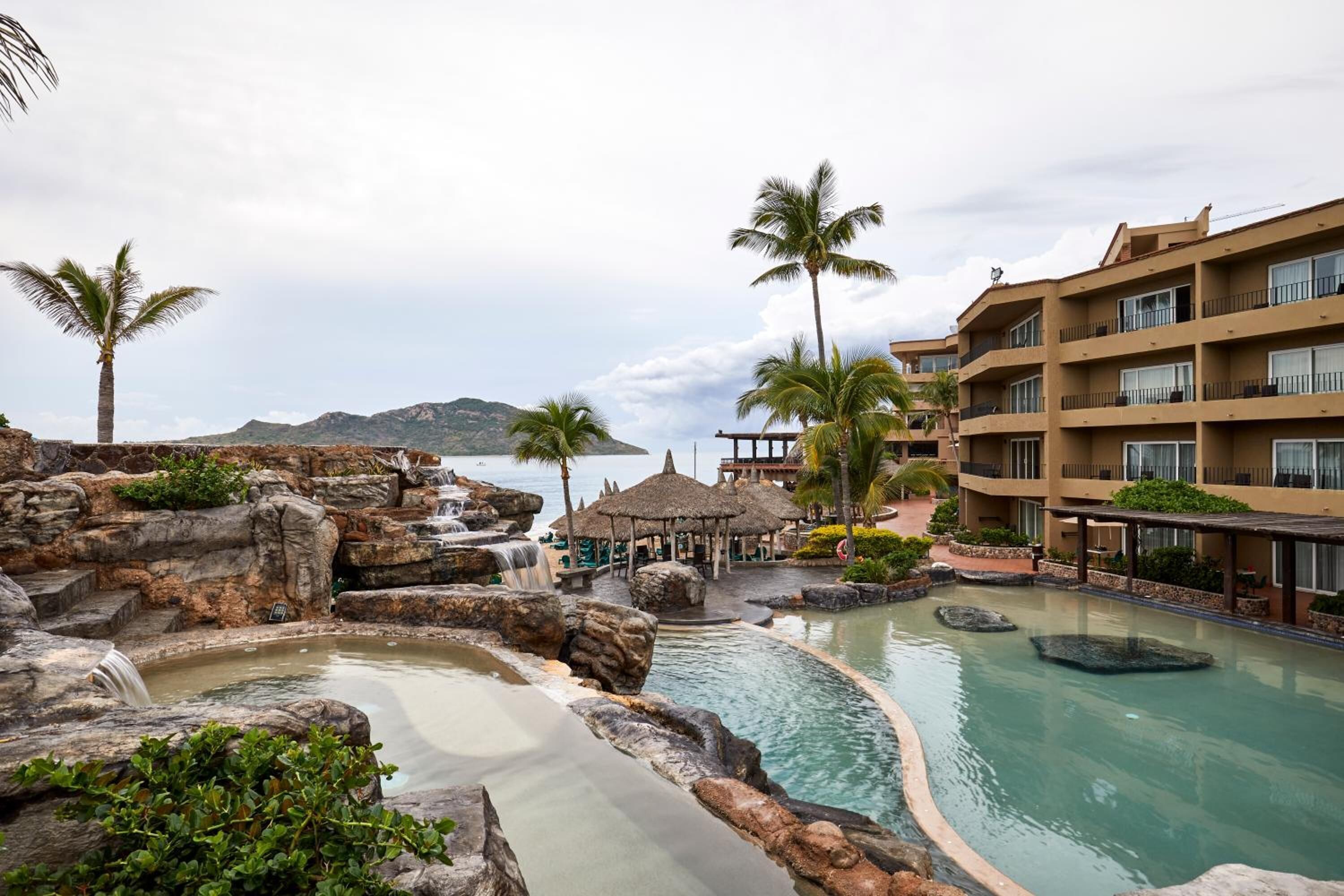Un resort con zona de piscina rodeada de rocas y palmeras, una cascada y un edificio con balcones.