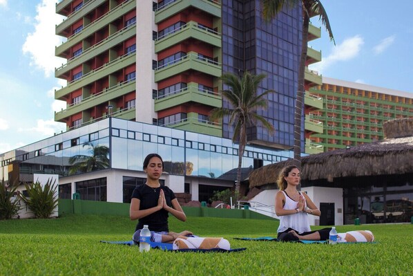 Two individuals meditating on a grassy lawn with a modern building and a thatched-roof structure in the background.