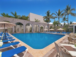 A swimming pool with blue lounge chairs and umbrellas, surrounded by palm trees and a stone wall. - El Cid Granada Hotel (Mazatlán)