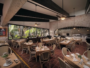 A restaurant with multiple tables and chairs, a stone wall, and a view of greenery through large windows. - El Cid Granada Hotel (Mazatlán)