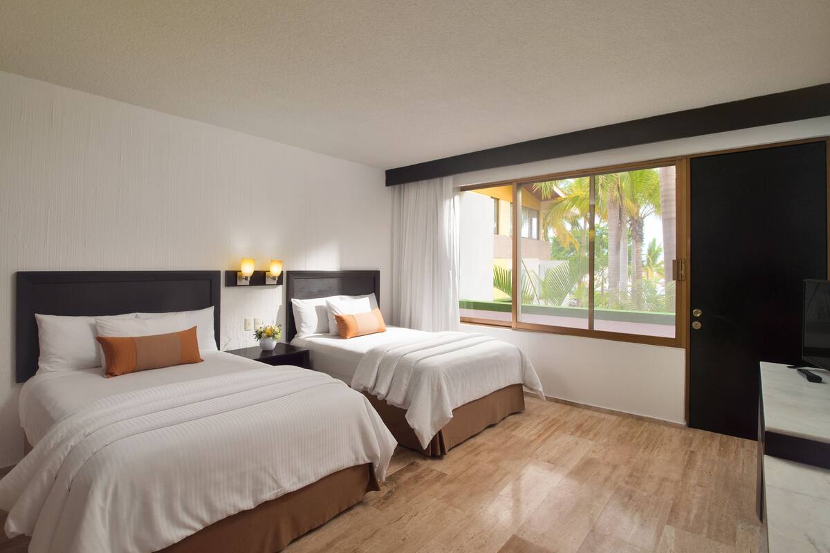 A hotel room with two beds, a desk, a TV, and a window with a view of palm trees.