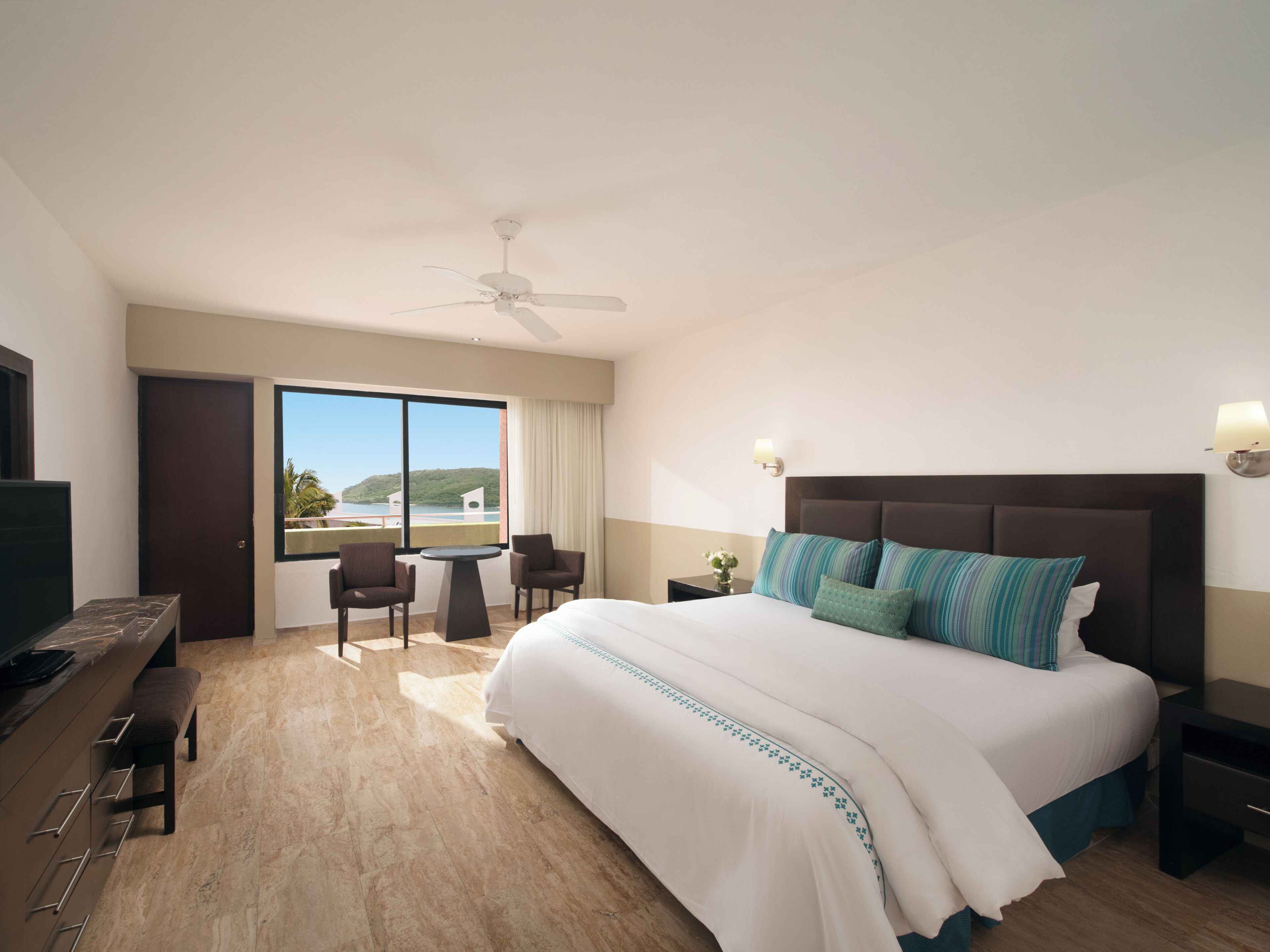 Une chambre d’hôtel avec un grand lit, un ventilateur de plafond, une télévision, un bureau avec deux chaises et une vue sur l’extérieur.