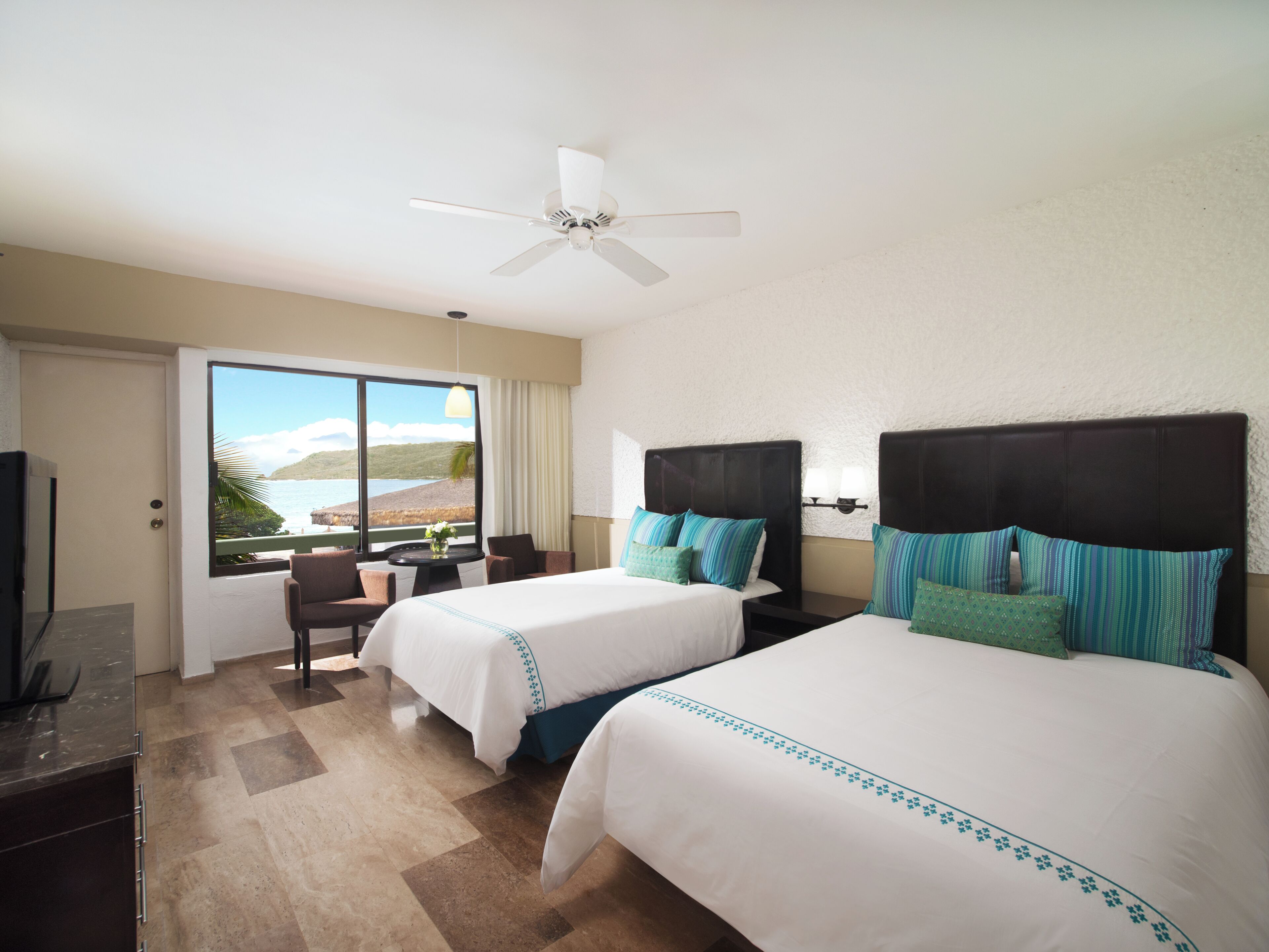A hotel room with two beds, a ceiling fan, a view of the ocean, a desk, and a chair.