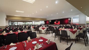 A large banquet hall with rows of tables set for dining, a stage with a red curtain, and a projector screen.