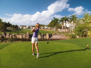 A golf course with a player in mid-swing, a putting green, and a backdrop of palm trees and buildings. - El Cid Castilla Beach Hotel (Mazatlán)