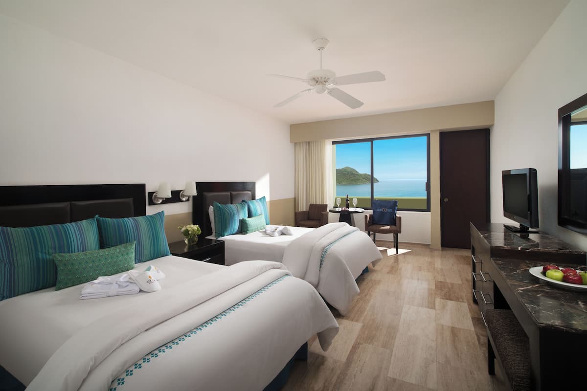 A hotel room with two beds, a desk with a television, a view of the sea, and a ceiling fan.