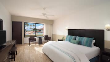 A hotel room with a bed, a desk, a chair, a table, a ceiling fan, and a view of a pool and palm trees.