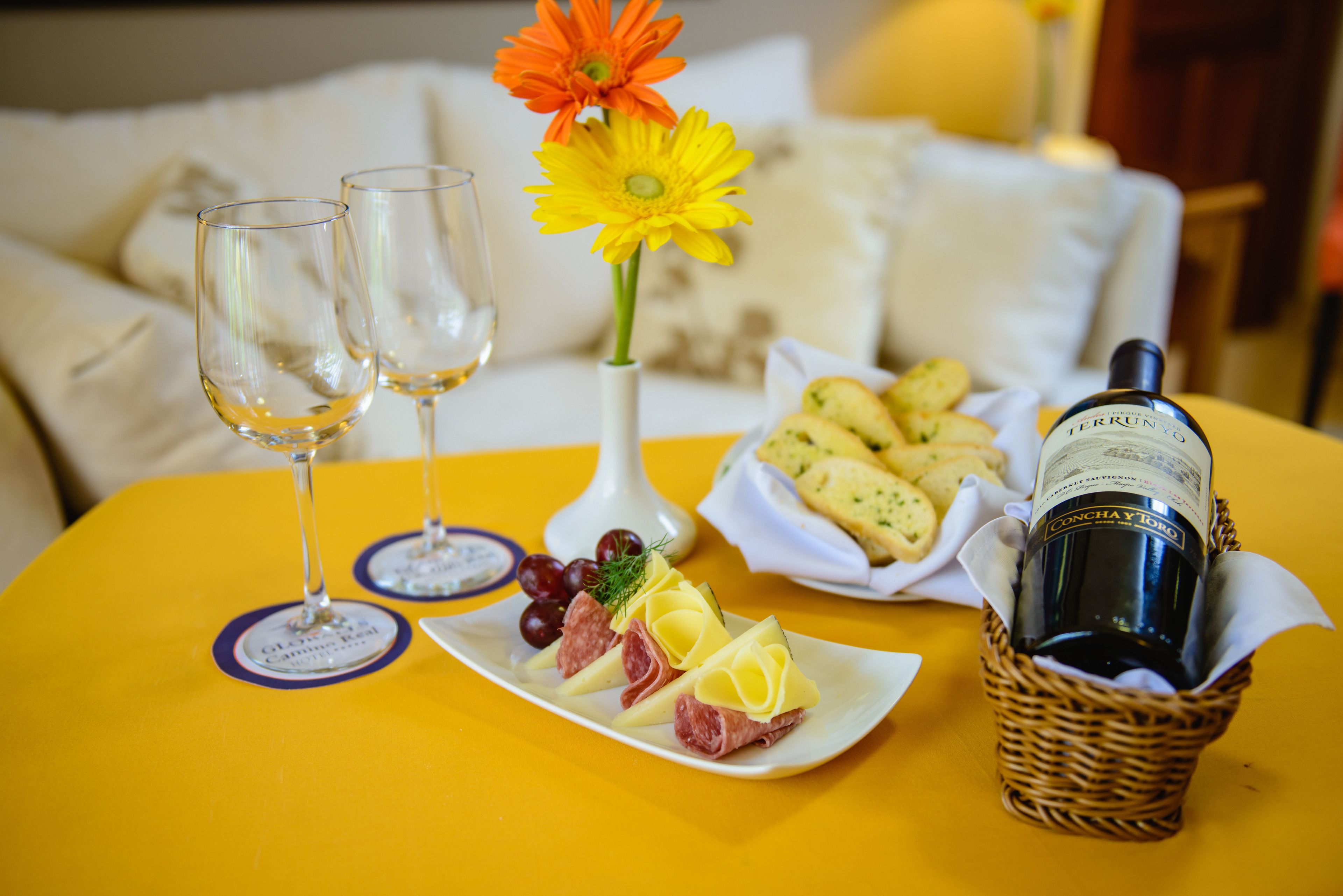 Uma mesa posta com dois copos de vinho branco, um prato de queijos e embutidos, uma cesta de biscoitos, uma garrafa de vinho e um vaso com flores.